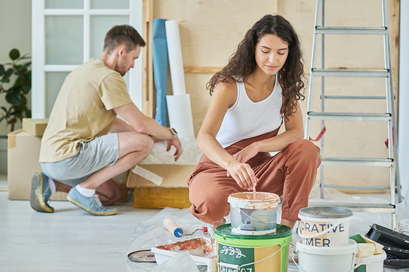 Young brunette woman mixing beige liquid paint in plastic bucket