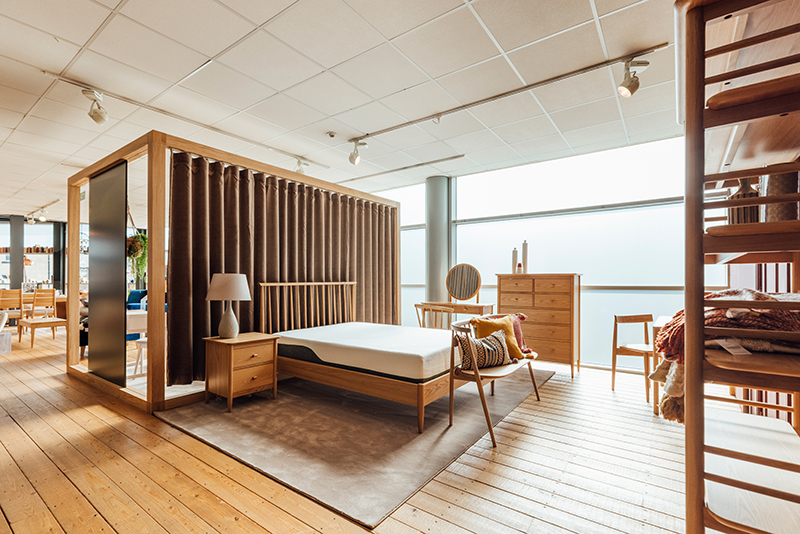 Wide angle image of a wooden showroom bed with a luscious mattress, light beams through the big windows, creating a clean effect across the image.