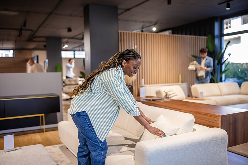 In a modern showroom, a woman carefully places a white pillow onto a chic sofa, highlighting the inviting and stylish atmosphere of the furniture store.
